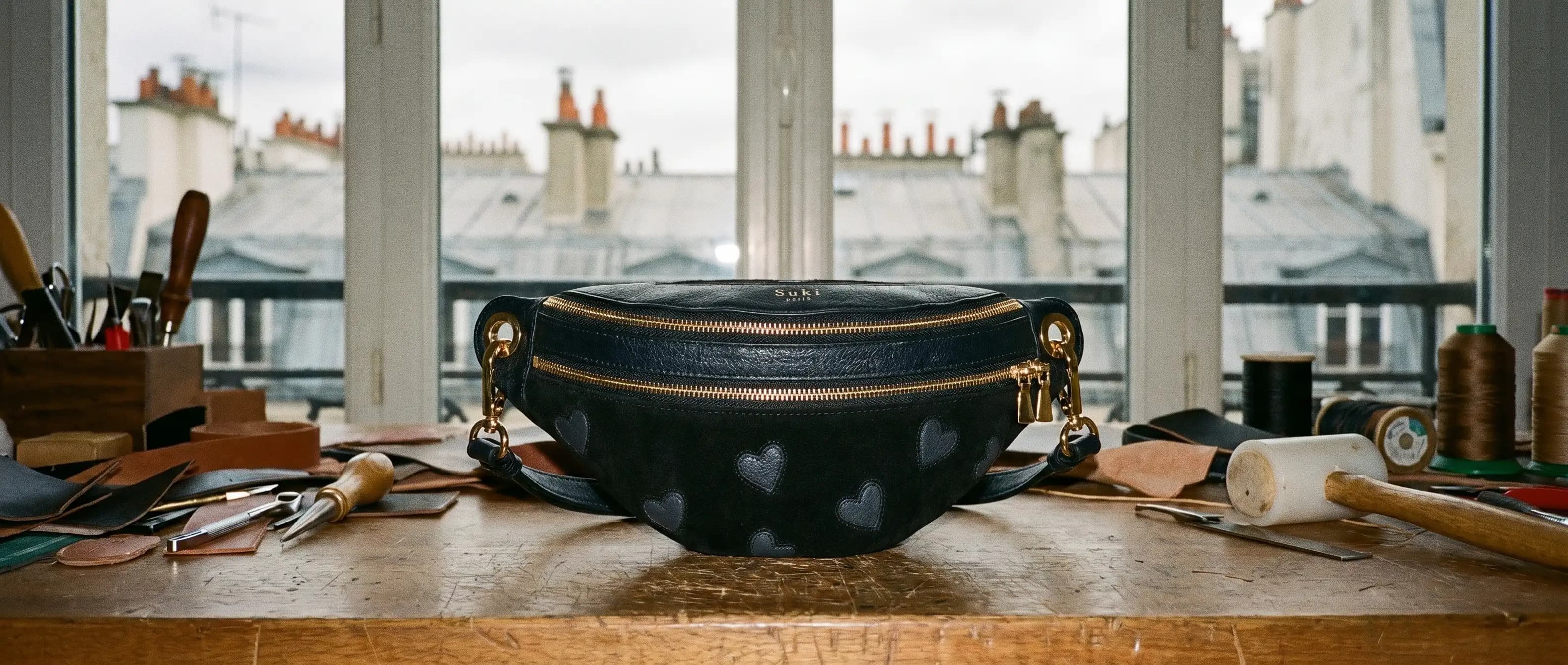 Établi de l'atelier Suki devant les toits de Paris, avec un sac banane en cuir noir à cœurs incrustés